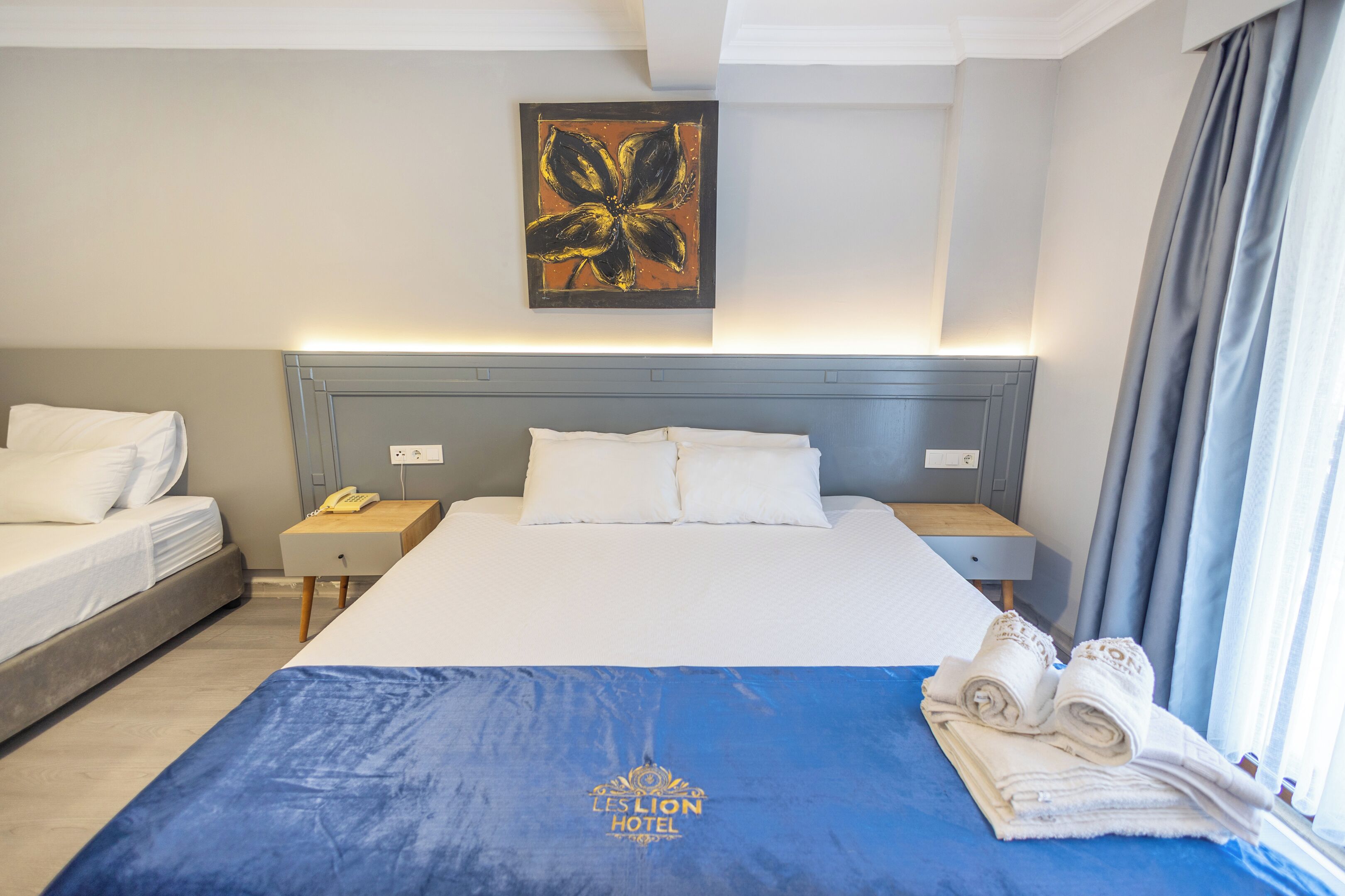Habitación de hotel con una cama grande, otra más pequeña, una mesita de noche con lámpara, un cuadro en la pared y una colcha azul con el logotipo del hotel.
