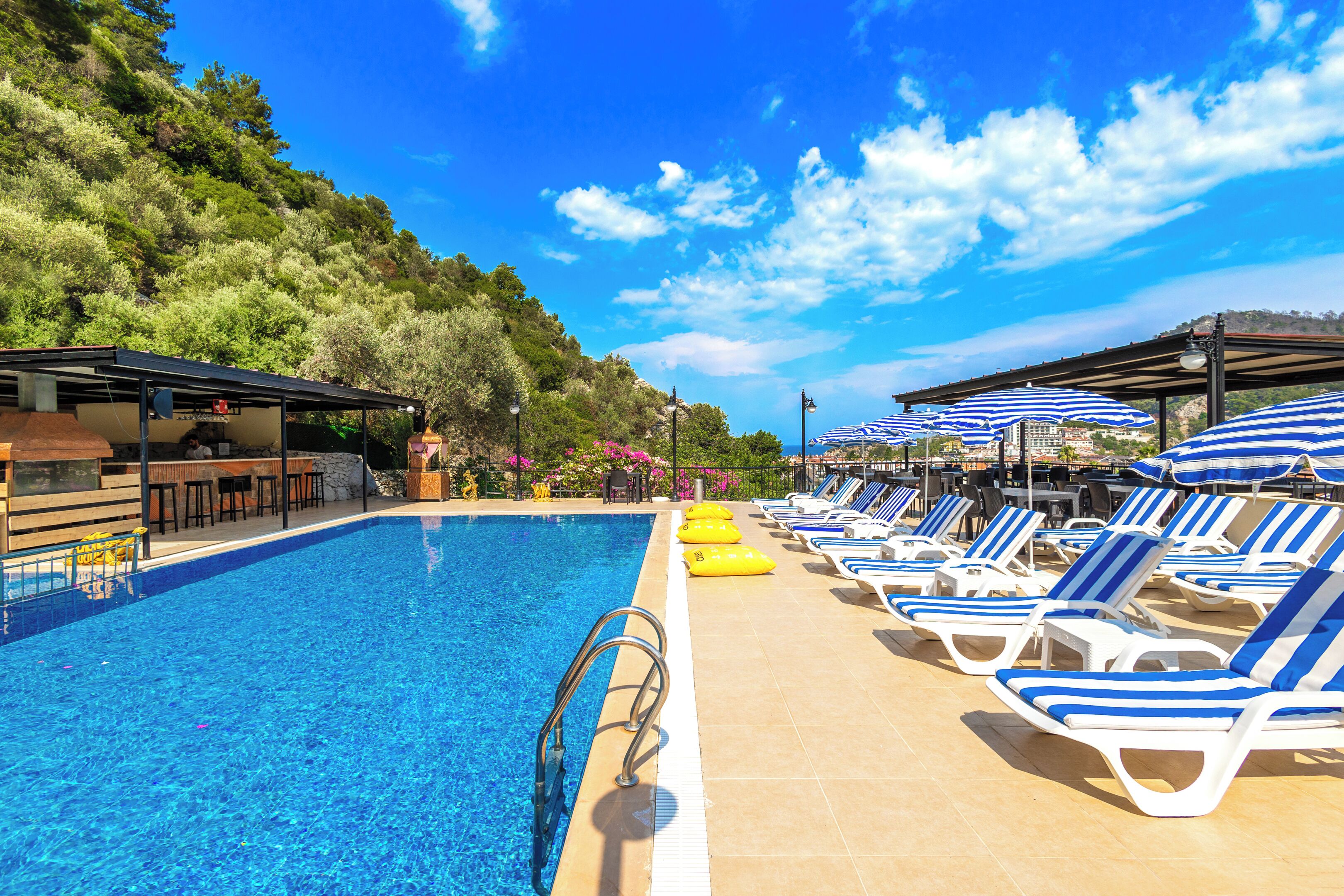 A swimming pool with lounge chairs and umbrellas, surrounded by greenery and a clear sky.
