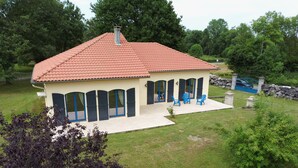 A two-story house with a red-tiled roof, a covered patio with blue chairs, and a stone wall in the background. - At the foot of the mountains: villa des nestes (Saint-Paul)
