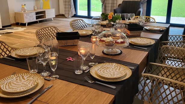 A dining area with a large table set for six, surrounded by wicker chairs. A television is mounted on the wall, and a sofa is visible in the background. - At the foot of the mountains: villa des nestes (Saint-Paul)