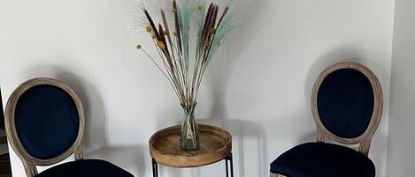Two upholstered chairs with dark cushions, a small wooden table with a vase of dried plants, and a framed picture on the wall.