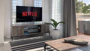 Uma sala de estar moderna com uma TV de tela plana grande exibindo o logotipo da Netflix, um rack de madeira, uma planta em vaso e uma área de jantar com mesa e cadeiras.