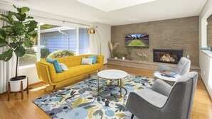 A modern living room with a yellow sofa, grey armchair, and a round coffee table. A large window provides natural light, and a fireplace is visible in the background. - Mid century modern retreat w/ expansive garden and central A/C (Lake Forest Park)