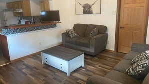 A living room with a sofa, a coffee table, and a TV. There is a kitchen area with cabinets and a TV. A door and a wall with a bull picture are also visible. - Experience Sturgatory 1BR by Main & Junction (Sturgis)
