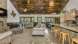 A spacious living area with a stone fireplace, a wooden ceiling, and a large window overlooking greenery. - Prickly Pear, Pool, Golf Course Home, @ Frio River Vacation Rentals (Concan)