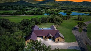 A large, modern house with a metal roof, surrounded by lush greenery and a lake. - Prickly Pear, Pool, Golf Course Home, @ Frio River Vacation Rentals (Concan)