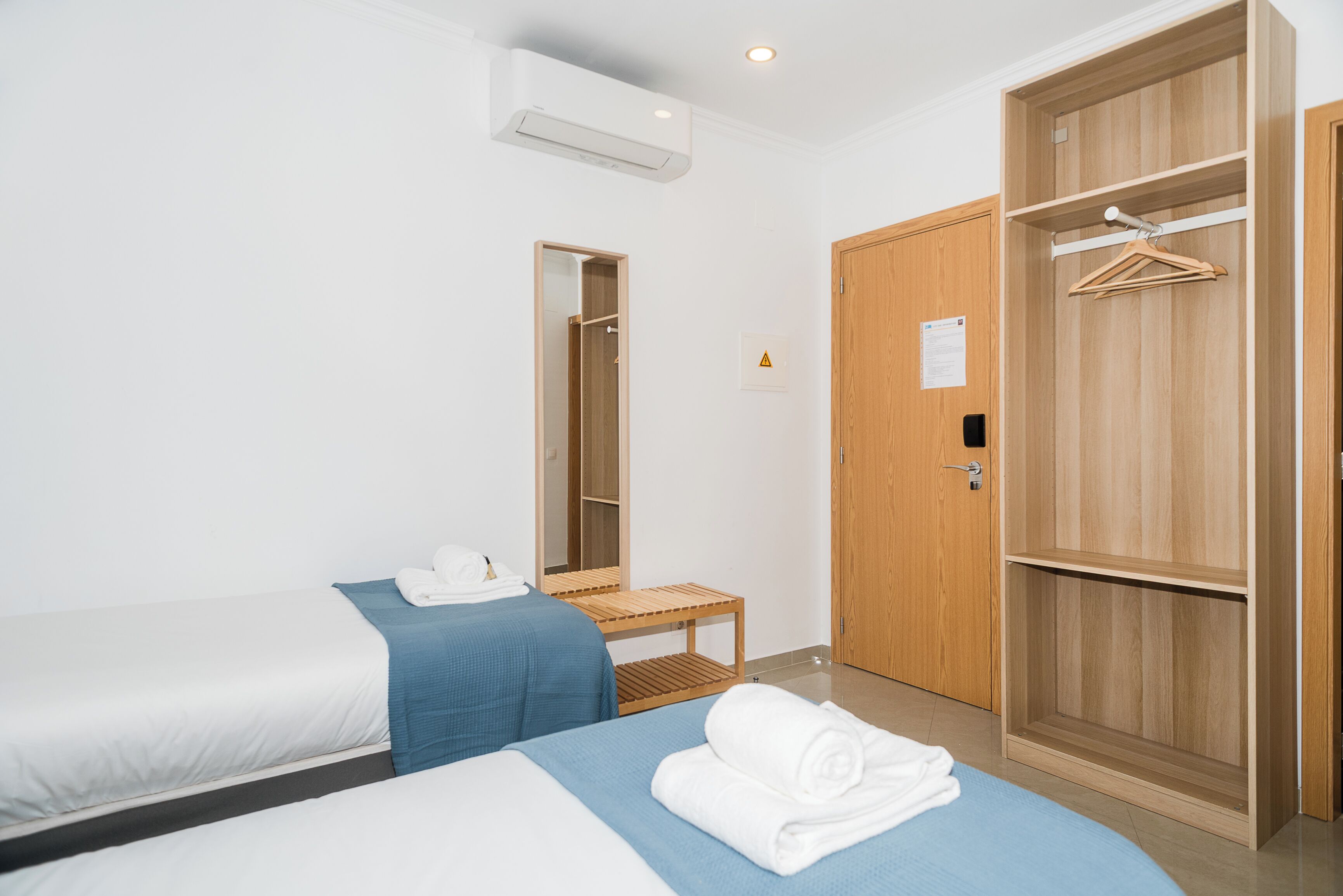 Une chambre d’hôtel avec deux lits, une armoire en bois, un miroir, une petite table et un appareil de climatisation.