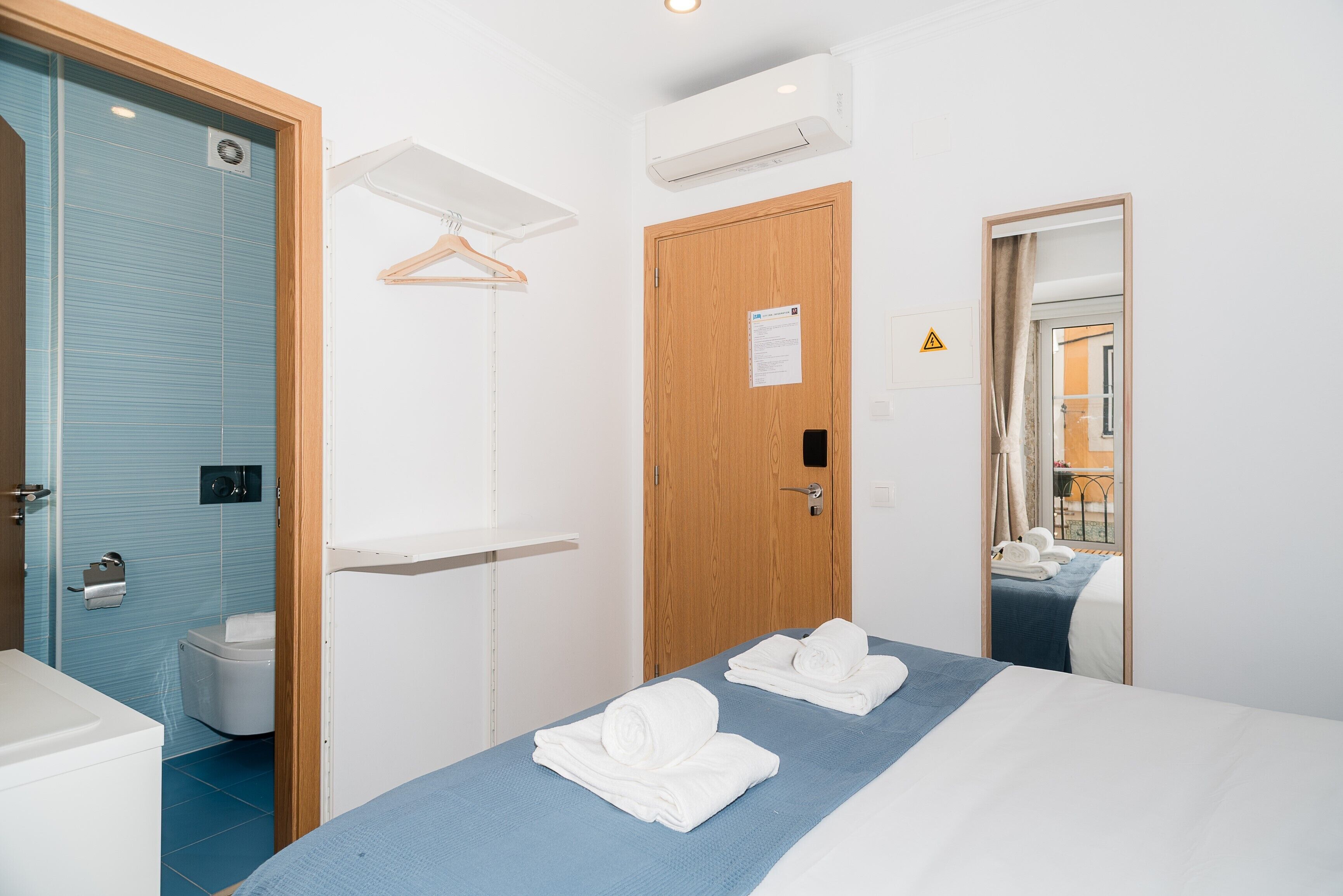 A hotel room with a bed, a wooden door, a towel rack, a mirror, and an air conditioning unit.