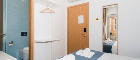 A hotel room with a bed, a wooden door, a towel rack, a mirror, and an air conditioning unit.