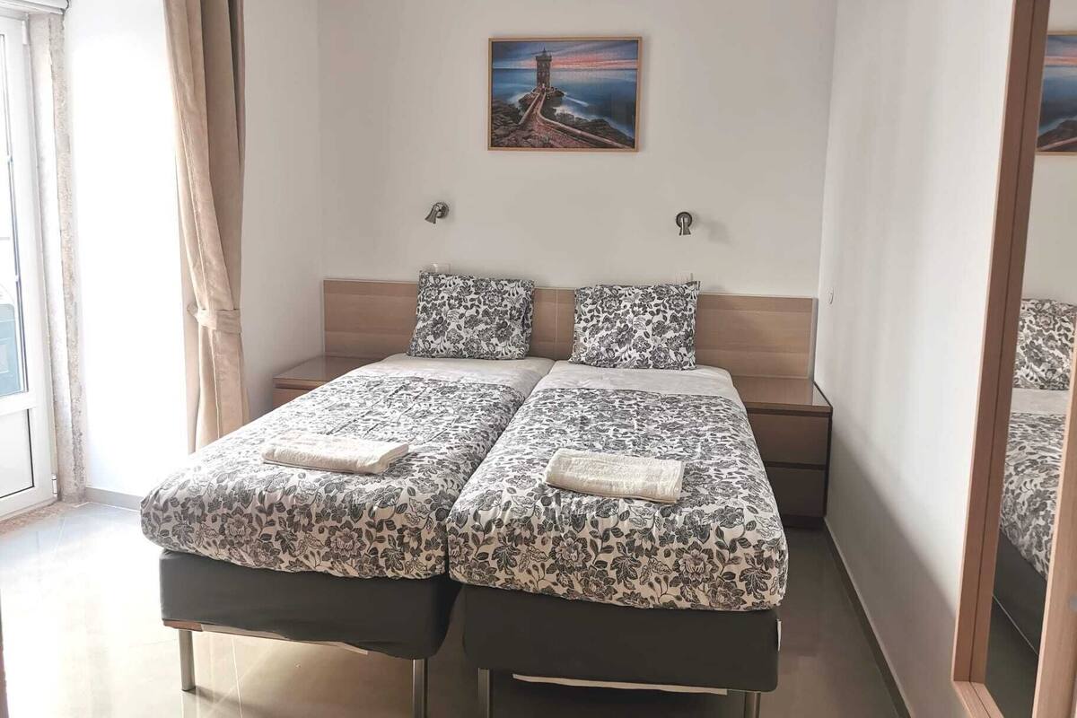 Una cama doble con ropa de cama floral, una mesita de noche, una lámpara de pared y un cuadro de una escena costera.