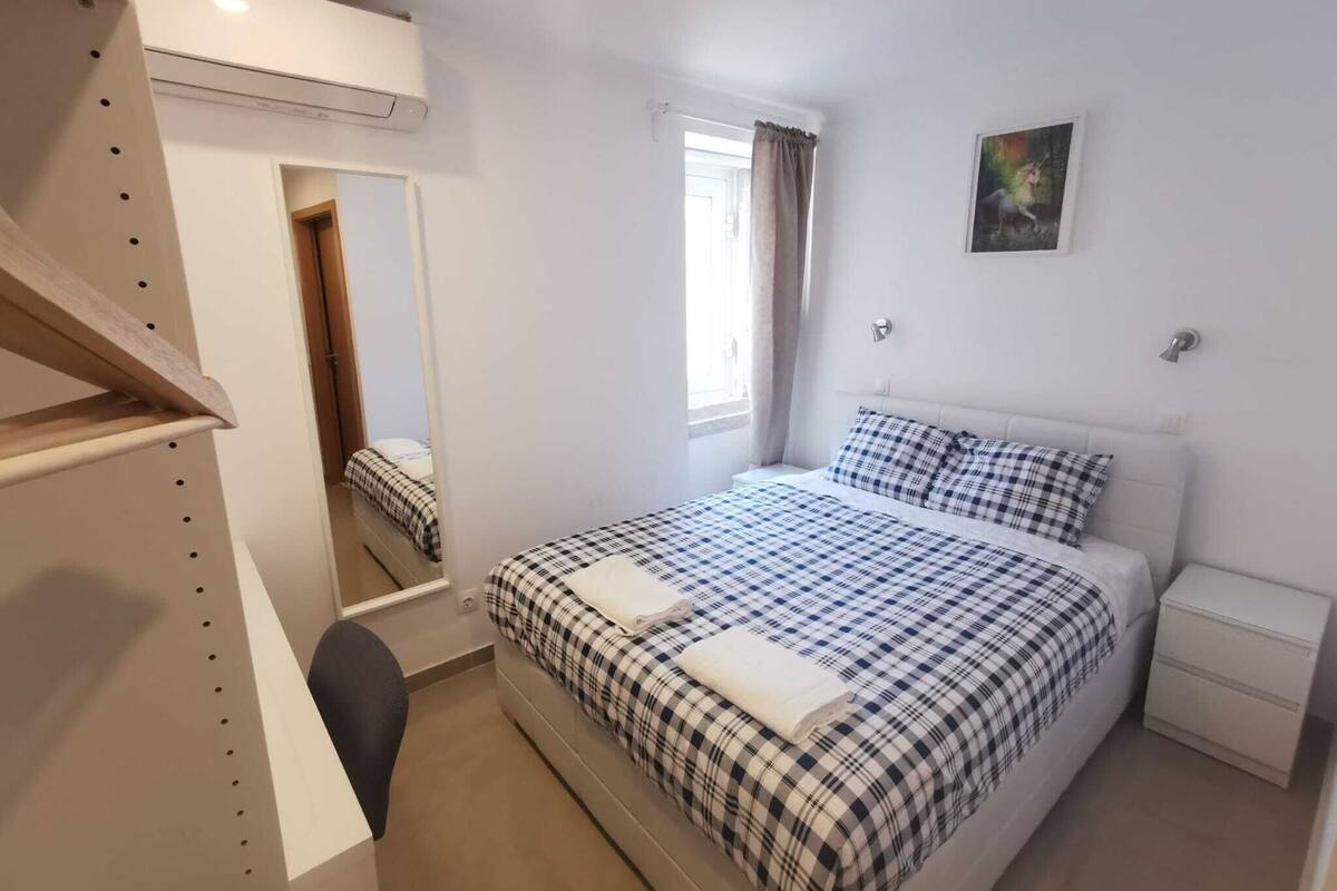 Un dormitorio con cama, mesita de noche, silla, armario, ventana con cortinas y un cuadro en la pared.