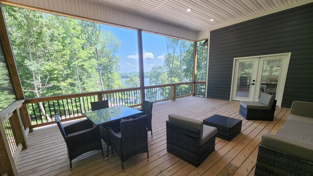 A covered deck with a view of a lake, furnished with wicker chairs and a glass-top table.