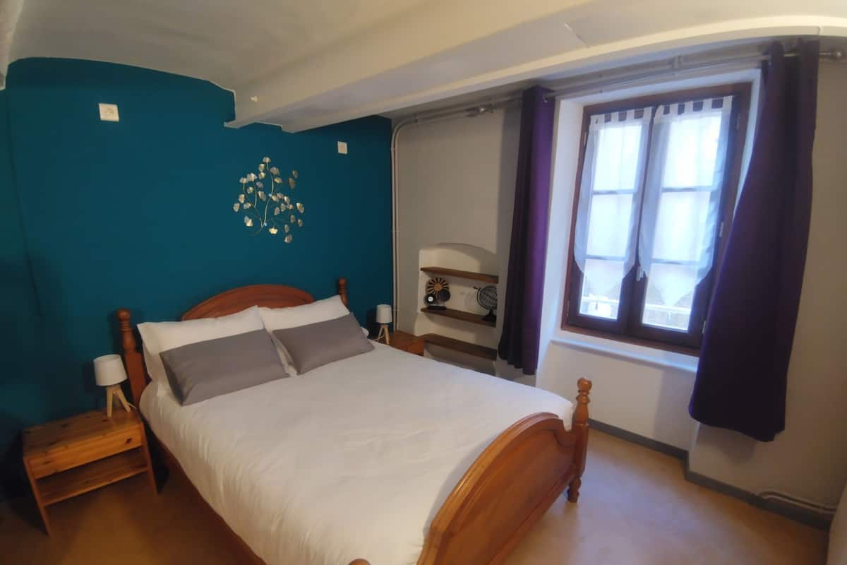 Une chambre à coucher avec un lit en bois, une table de chevet, une horloge murale et une fenêtre avec des rideaux.