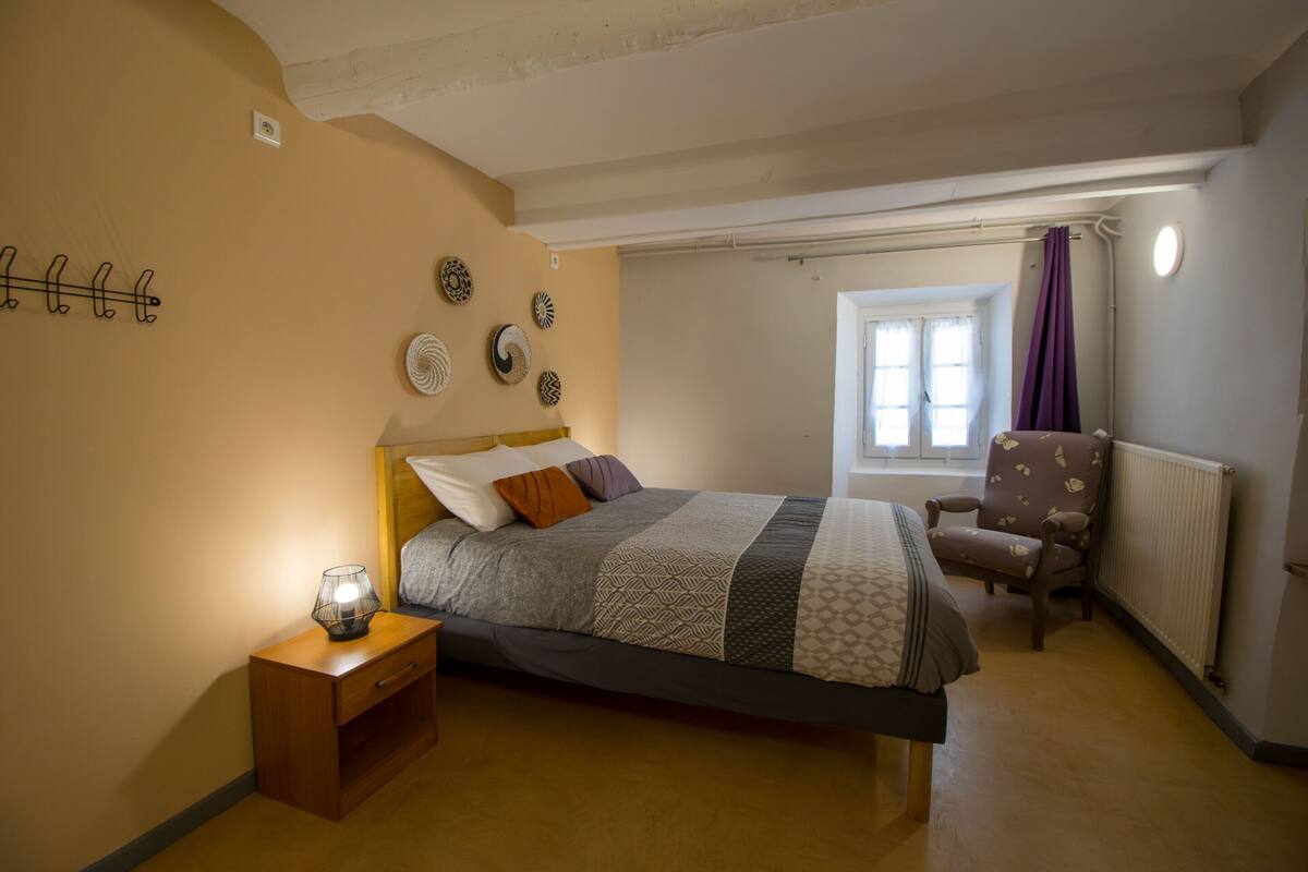 Une chambre à coucher avec un lit, une table de chevet, une chaise et une fenêtre avec des rideaux.