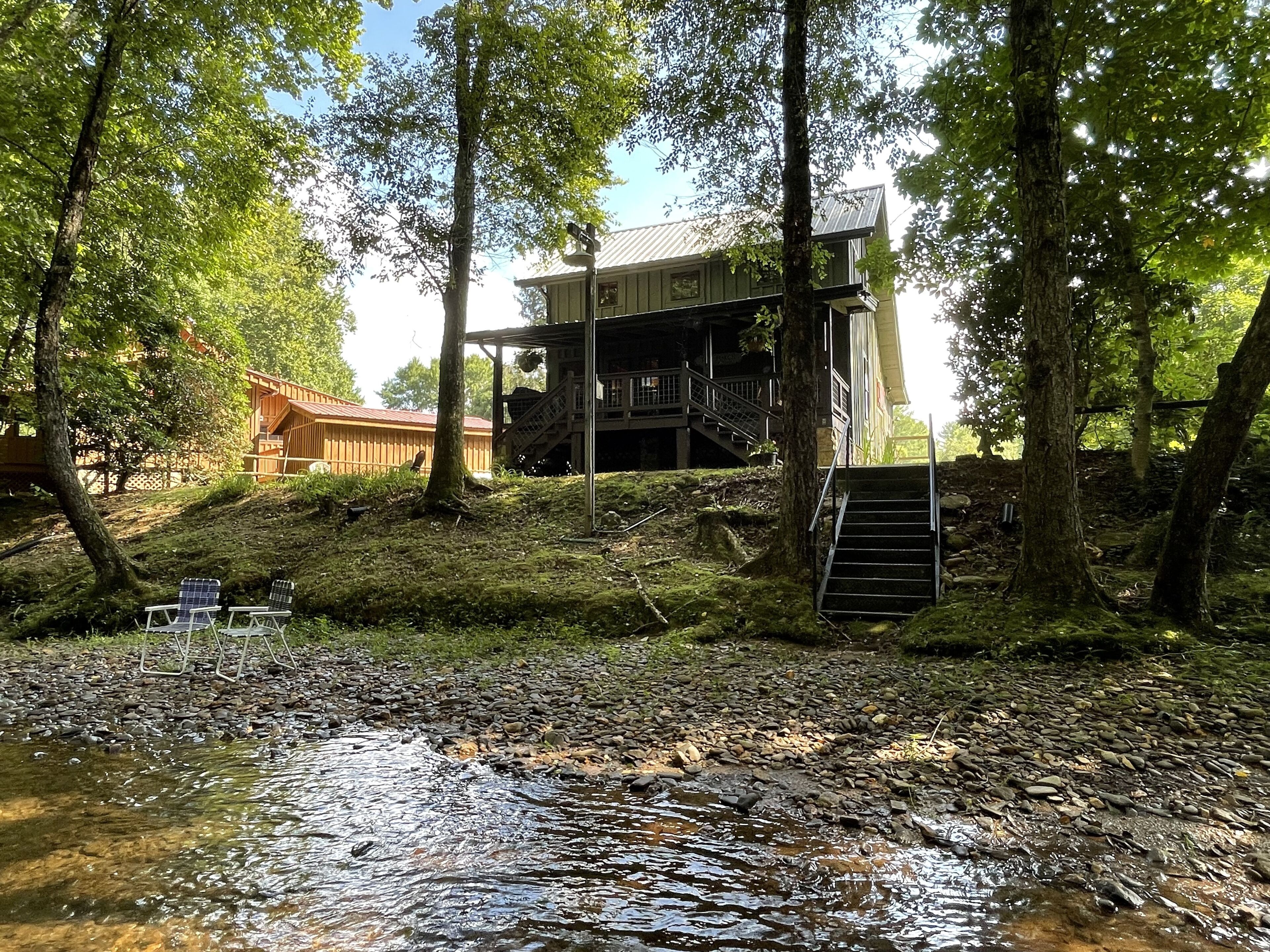 A cabin with a wooden deck, a staircase leading to a second level, and a stream in the foreground.
