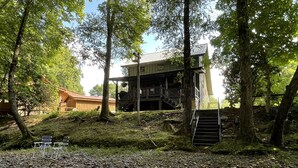 Una cabaña con terraza de madera, una escalera que lleva a un segundo nivel y un arroyo al frente.