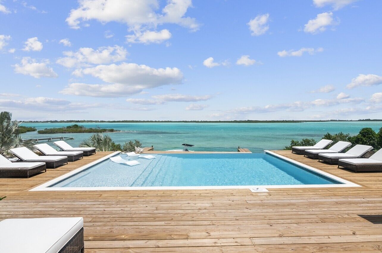 Ein Pool mit Liegestühlen, eine Holzterrasse und Blick auf das Meer und Inseln.