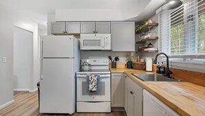 A compact kitchen with white appliances, a wooden countertop, and a window with blinds. - Family Friendly & Cheerful Blue Jay Ranch House close to KSU stadium  (Manhattan)