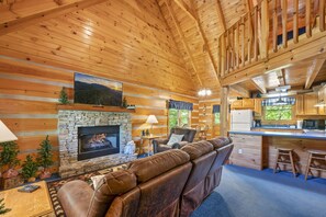 A log cabin interior with a stone fireplace, leather sofa, dining area, and kitchen.
