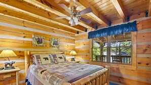 Ein Schlafzimmer einer Holzhütte mit Bett, zwei Nachttischlampen, Deckenventilator und Fenster mit Blick ins Freie.