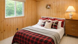A bedroom with a wooden paneled wall, a bed with red and white bedding, a nightstand with a lamp, and a window with a view of trees.