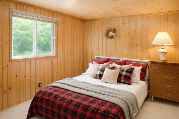 A bedroom with a wooden paneled wall, a bed with red and white bedding, a nightstand with a lamp, and a window with a view of trees.
