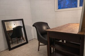 A room with a wooden table and a black chair, a mirror reflecting part of the room, and a window with a view of trees.