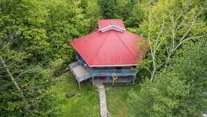 A cabin with a red roof, surrounded by dense green forest, accessible by a wooden deck.