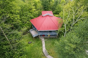 A cabin with a red roof, surrounded by dense green forest, accessible by a wooden deck.