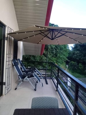 A balcony with two reclining chairs, a covered area with string lights, and a view of greenery.