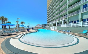 Un espace piscine d’hôtel avec des chaises longues et des parasols, une piscine à l’eau d’un bleu limpide et un bâtiment doté de balcons.