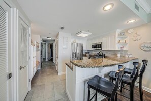 A modern kitchen with white cabinetry, granite countertops, and a central island with bar stools. - FREE Perks! Boardwalk 1907, King Master, New Walk In Shower & Heated Pool! (Panama City Beach)