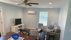 A modern living room with a dining area, a ceiling fan, a television on a cabinet, a window with blinds, and a gray sofa with patterned cushions.