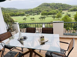 A glass-top table with four chairs, a bottle, and a tissue box on a balcony overlooking a green landscape with trees and hills. - FLAT Surrounded by nature, with pool, and beach nearby (ES MERCADAL MENORCA)