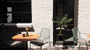 Un pequeño espacio al aire libre con dos mesas redondas, cuatro sillas, una planta en maceta y una ventana con persianas.