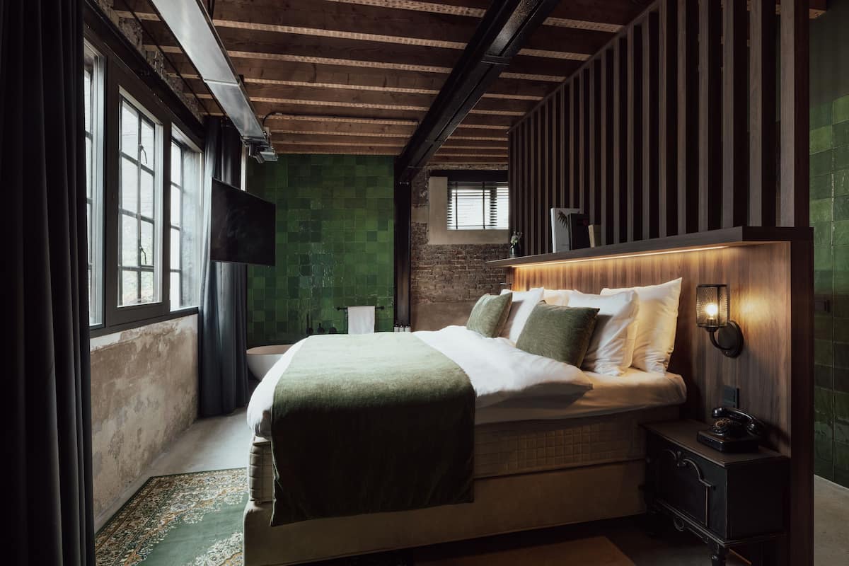 A bedroom with a large bed, a nightstand, a wall-mounted lamp, and a window with curtains.