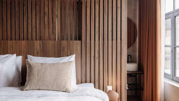 Un dormitorio con cabecera de madera con listones, cama con ropa de cama blanca, mesita de noche redonda color naranja, y vista a un baño a través de una ventana.
