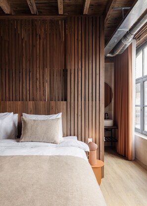 A bedroom with a wooden slat headboard, a bed with white linens, a round orange side table, and a view of a bathroom through a window. - Cousins Boutique Hotel (Maastricht)