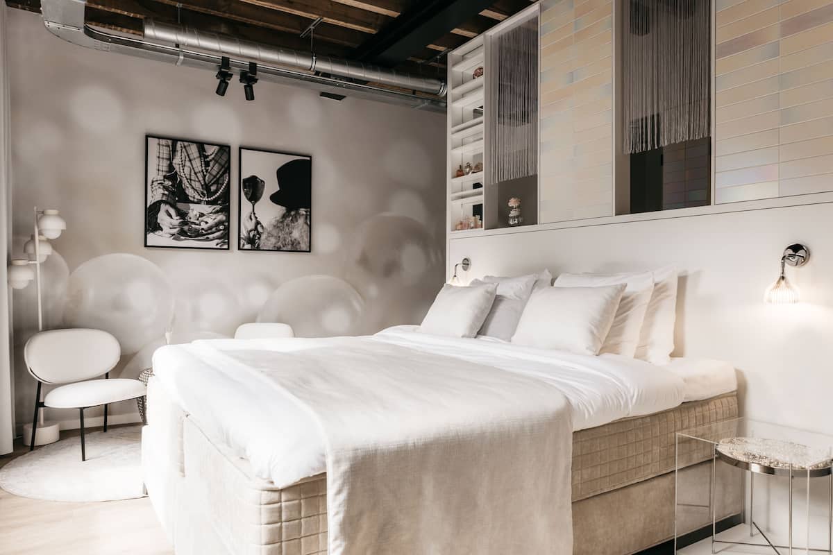 A modern bedroom with a large bed, a side table, a chair, and a wall-mounted shelf with decorative items.