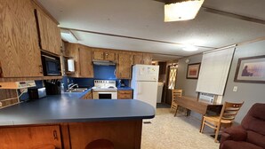 A compact kitchen with wooden cabinets, a central island with stools, a dining area with a table and chairs, and a refrigerator.