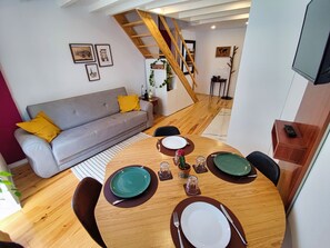 A modern living room with a grey sofa, wooden dining table set for two, and a staircase leading to an upper level.