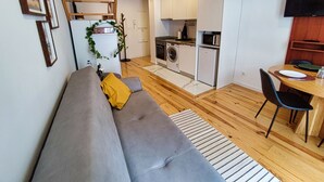 Ein kompakter Wohnbereich mit grauem Sofa, einem Esstisch mit Stühlen, einer Küchenzeile mit Geräten und einer Holztreppe.