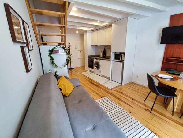 A compact living space with a grey sofa, a dining table with chairs, a kitchen area with appliances, and a wooden staircase.