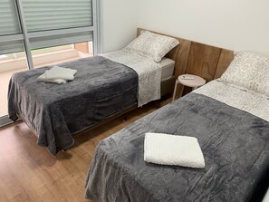 Two single beds with floral-patterned bedspreads, a small table, and a large window with horizontal blinds.