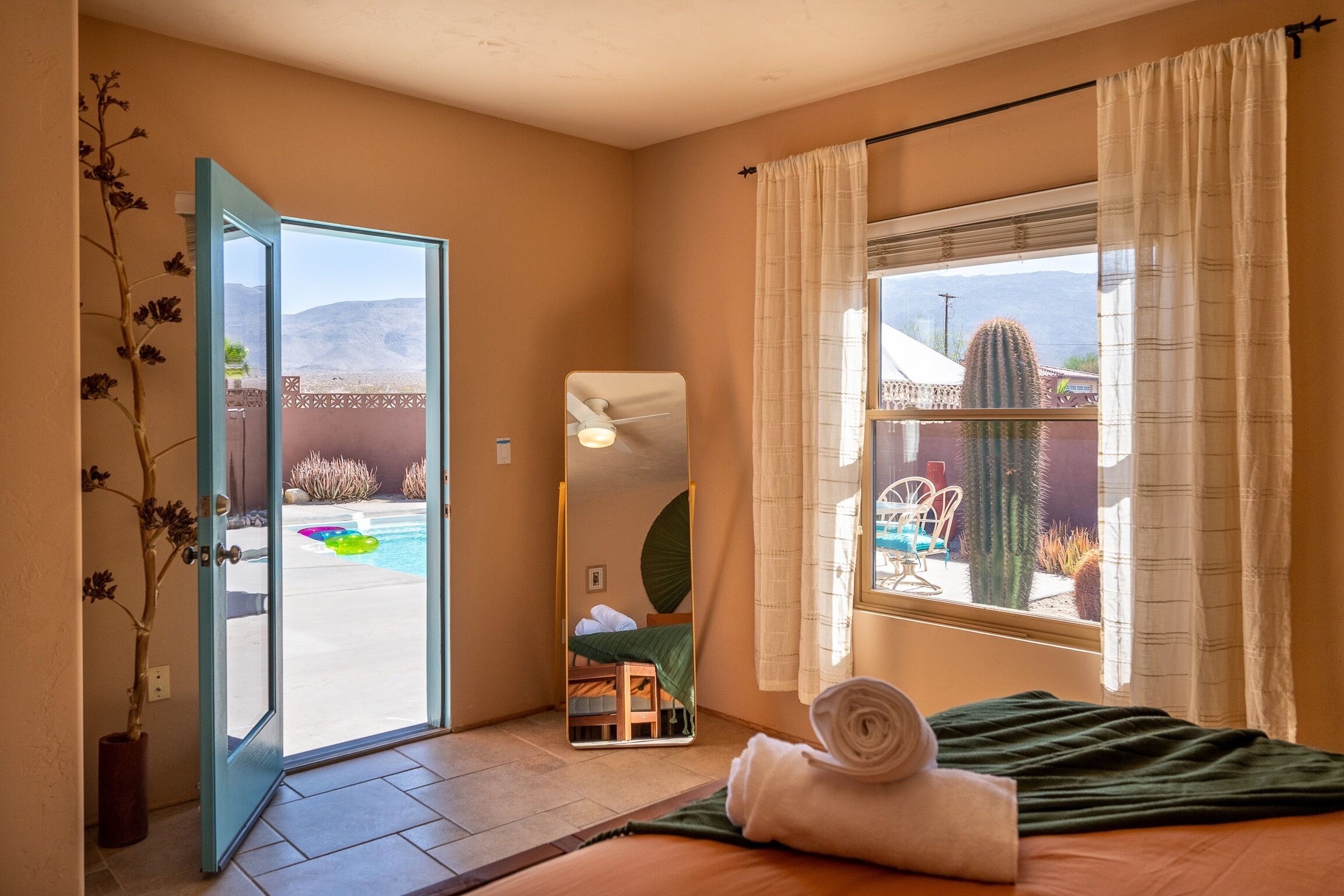 Una habitación con una cama, una ventana con cortinas, un espejo y una puerta que da a una zona de piscina.
