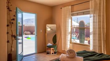 Una habitación con una cama, una ventana con cortinas, un espejo y una puerta que da a una zona de piscina.