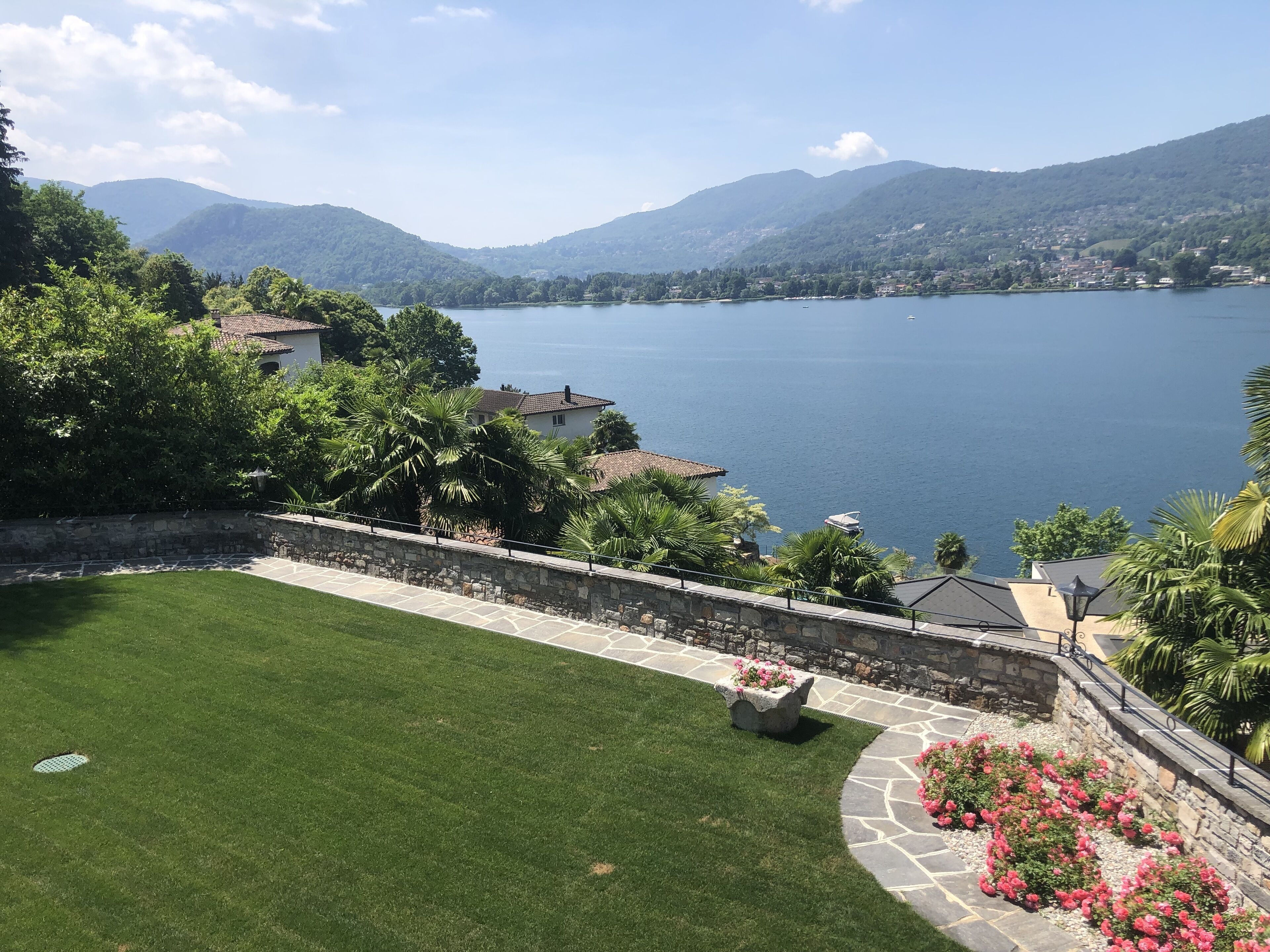 Vista de un lago con montañas al fondo, un césped cuidado y un muro de piedra con macizos de flores.