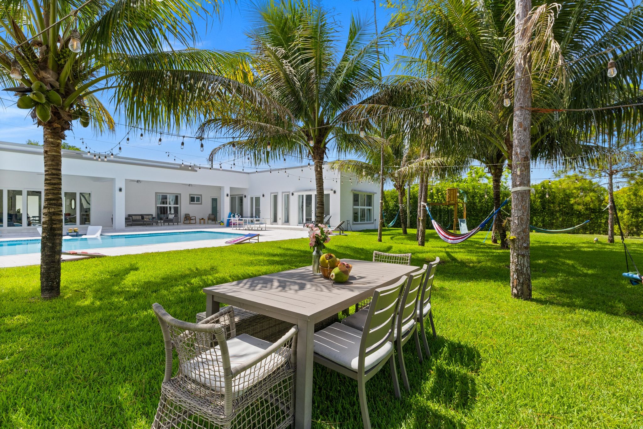 Área de piscina com mesa e cadeiras, cercada por palmeiras e um prédio branco.