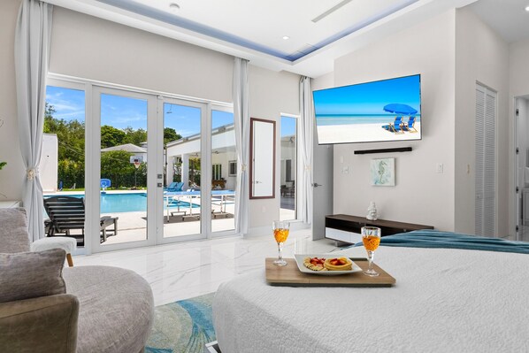 A modern living room with a large TV, a dining area with a tray of food, and a view of a pool and beach.