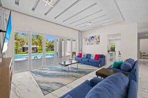 A modern living room with a blue sofa, a coffee table, and a view of a swimming pool through glass doors. - The White House of Miami, Perfect for Your Family (Princeton)